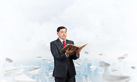 Serious businessman holding open book. Man in business suit standing on cloudy cityscape background with flying paper planes. Lawyer reading legal regulation book. Business accounting and consultingの写真素材