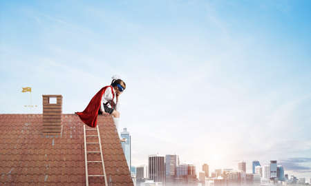 Little sad child in mask and cape sitting on roof edge and looking down. Mixed mediaの写真素材