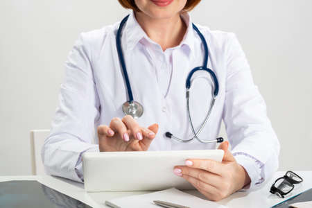 Close up female doctor hands using tablet computer. Family physician in white coat with stethoscope sitting at desk. Professional medical diagnosis and treatment in clinic. Medical online applicationの写真素材