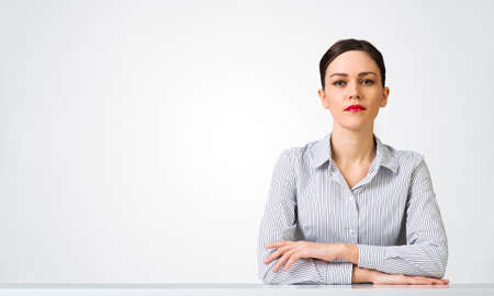Confident businesswoman sitting at desk with folded hands. Young caucasian female student has serious facial expression. Portrait of girl wears shirt on white background. People emotion and expressionの写真素材