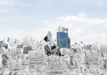 Young businessman looking away while sitting on pile of documents among flying letters with cityscape on background. Mixed media.の写真素材