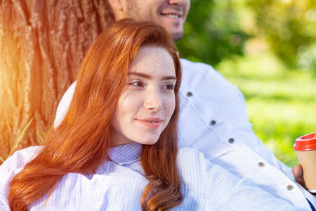 Young couple relaxing under tree in summer park on sunny day. Happy couple in love spend time outdoors together. Handsome man and pretty redhead girl enjoying each other. Romantic relationships.の写真素材