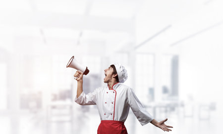 Young chef shouting loudly into megaphone. Emotional caucasian chef in white hat and red apron on light blurred background. Restaurant advertisement, food retail campaign announcement.の写真素材