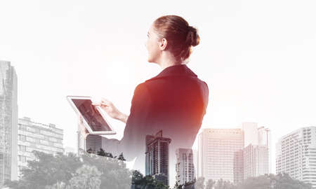 Young businesswoman using tablet computer. Double exposure concept with modern cityscape and beautiful woman in business suit. Digital technology in property management and development company.の写真素材