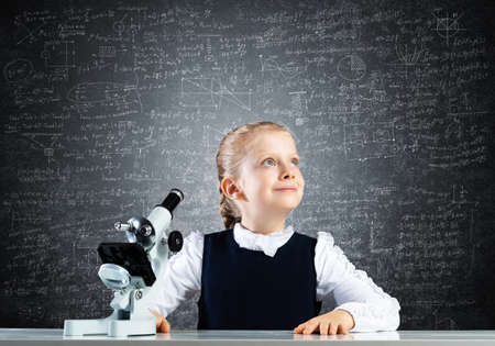 Little girl scientist with microscope on chalkboard background. Research and discovery concept with copy space. Elementary science class in modern school. Schoolgirl in schoolwear sitting at desk.の写真素材
