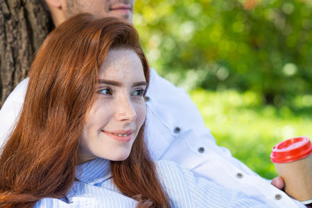 Young couple relaxing with coffee under tree in park on sunny day. Happy couple in love spend time outdoors together. Handsome man and pretty redhead girl sitting on green grass leaning against treeの写真素材