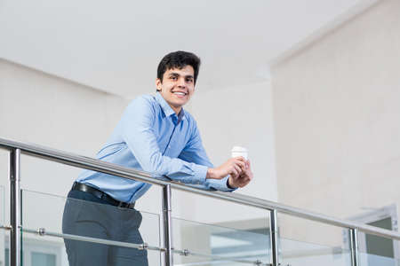 a young businessman leans on a railing. holds a glass of coffee and smilesの写真素材