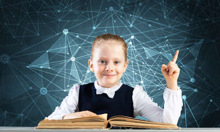 Smiling little girl sitting at desk with open book. Research and education in school. Happy schoolgirl in school wear finger pointing up. Clever girl reading book on deep blue plexus backgroundの写真素材