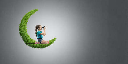 Cute kid girl sitting on green moon and looking in binocularsの写真素材
