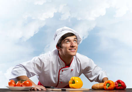 Young smiling chef standing near cooking table with fresh vegetables. Pleased chef in white hat and...