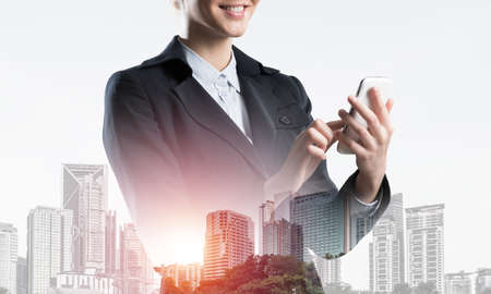 Young businesswoman using mobile phone. Double exposure concept with modern downtown architecture and beautiful woman in business suit. Digital technology in property management and development.の写真素材
