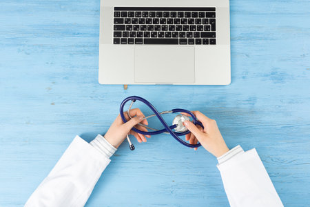Top view of doctor hands with stethoscope. Therapist in white coat sitting at blue wooden desk with laptop. Examination and consultation in clinic. Medical application and online healthcare services.の写真素材