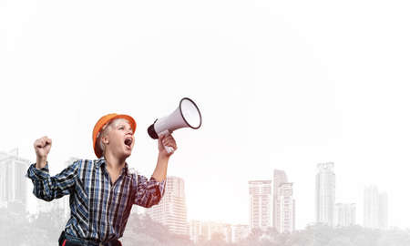 Expressive woman in safety helmet shouting into megaphone. Portrait of young emotional construction worker with wide open mouth on foggy city background. News announcement and advertisement.の写真素材