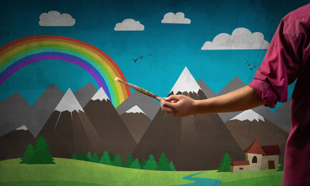 Close up artist hand holding paintbrush. Painter in shirt standing on background colorful picture. Summer landscape with mountains, blue sky and rainbow artwork. Creative hobby and profession.の写真素材