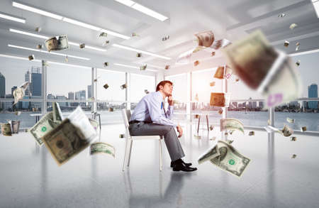 young businessman sits on an office chair. thoughtful and looking up. documents fly aroundの写真素材