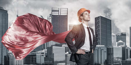 Confident engineer man in helmet and red cape against modern cityscape backgroundの写真素材