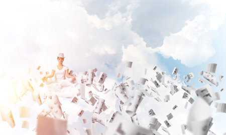 Woman in white clothing keeping eyes closed and looking concentrated while meditating among flying papers in the air with cloudy skyscape on background.の写真素材