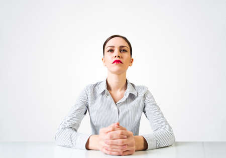 Confident businesswoman sitting at desk with folded hands. Young caucasian female student has serious facial expression. Portrait of girl wears shirt on white background. People emotion and expressionの写真素材