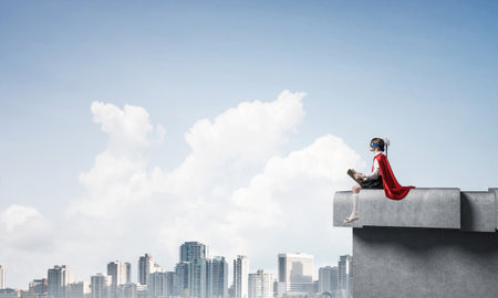 Little child in mask and cape sitting on building roof and reading . Mixed mediaの写真素材