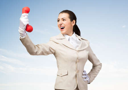 Expressive woman shouting into red handset phone. Operator in business suit standing with cellphone on blue sky background. Business assistance and consultation. Customer service support conceptの写真素材