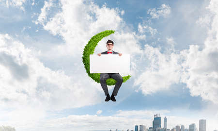 a young businessman with a poster sits on the green moon. urban landscape in the background.の写真素材