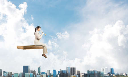 Elegant woman in business suit sitting on floating in air book. Musician with music brass instrument in hands. Attractive young woman with trumpet flying in blue sky above modern downtown.の写真素材