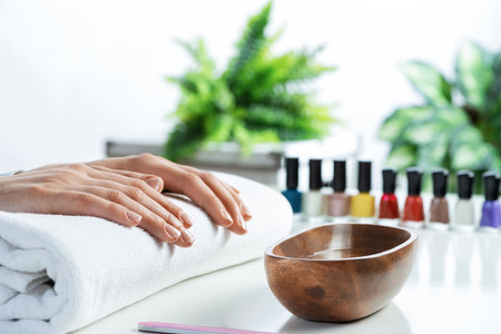 Female hands lying on white towel. Manicurist workspace with colorful nail polish bottles and wooden bowl with water. Female hand preparing for manicure. Professional nail care and beautician service.の写真素材