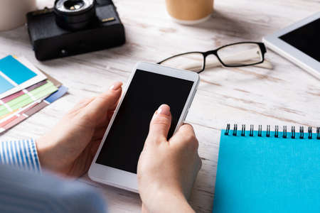 Blogger sitting at desk and holding smartphone. Mobile smart devices in business processes. Business occupation and innovation. Corporate office workplace with business woman at wooden desk.の写真素材