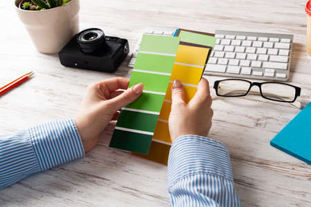 Interior designer holding color swatches at wooden desk. Office workplace with computer keyboard and camera. Brainstorming and creativity. Selection of color palette for web or product design.の写真素材