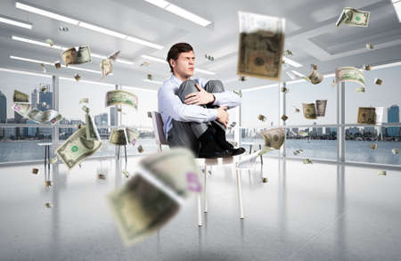 scared businessman sitting on a chair with his hands clasped his knees. money is flying around