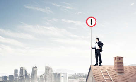 Young businessman with roadsign in hand standing on brick roof. Mixed mediaの写真素材
