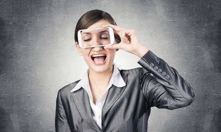 Portrait of pretty woman covering her eyes with smartphone. Happy businesswoman showing mobile phone with closed eyes on screen. Corporate businessperson on grey wall background. Mobile communicationの写真素材