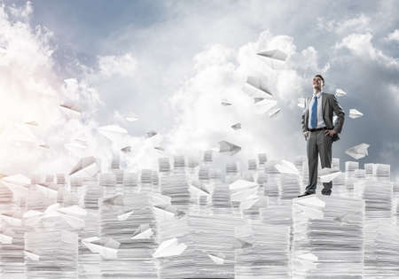 Confident businessman in suit standing among flying paper planes and looking away with cloudly skyscape on background. Mixed media.の写真素材