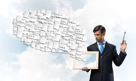 Portrait of young and successful businessman in black suit holding paintbrush in hands an looking at picture frameの写真素材