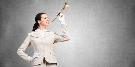 Beautiful woman playing trumpet brass. Young businesslady in white business suit and gloves posing with music instrument on grey wall background. Musician practicing and performing.の写真素材
