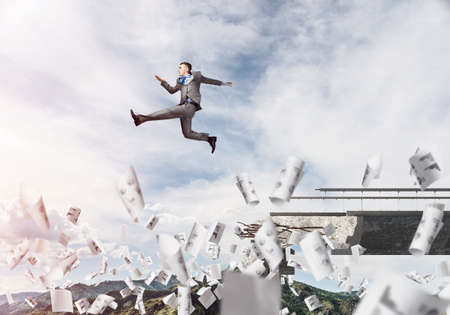 Businessman jumping over gap in bridge among flying papers as symbol of overcoming challenges....