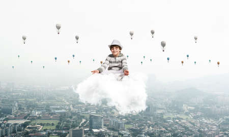 Young little boy keeping eyes closed and looking concentrated while meditating on cloud in the air with cityscape view and flying aerostats on background.の写真素材