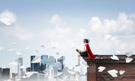 Little child in mask and cape sitting on building roof and reading . Mixed mediaの写真素材