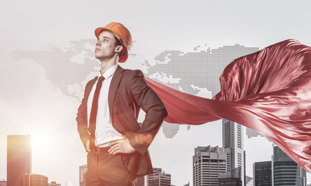 Confident engineer man in helmet and red cape against modern cityscape backgroundの写真素材