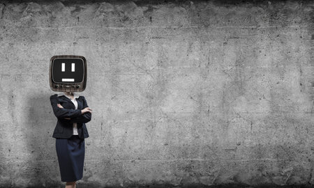 Business woman in suit with old TV instead of head keeping arms crossed while standing inside empty room with gray dark wall on background.の写真素材