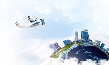 Businessman in aviator leather hat driving propeller plane. Traveling around the world by airplane. Funny man flying in small airplane in blue sky with clouds. Earth planet with modern cityscape.の写真素材