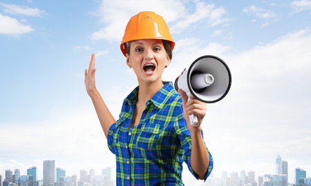 Expressive woman in safety helmet shouting into megaphone. Portrait of young emotional construction worker with wide open mouth on cityscape background. News announcement and advertisement.の写真素材