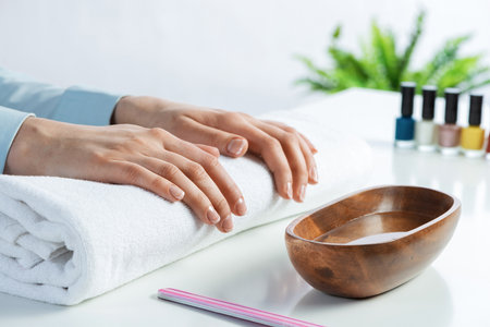 Female hands lying on white towel. Manicurist workspace with colorful nail polish bottles and wooden bowl with water. Female hand preparing for manicure. Professional nail care and beautician service.の写真素材
