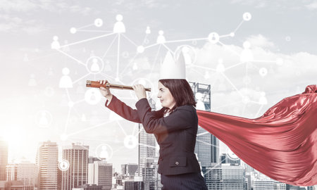 Young confident businesswoman wearing red cape against modern city background and looking in spyglassの写真素材