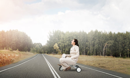 Beautiful happy woman riding childrens bicycle on asphalt road. Young employee in white business suit cycling small bike outdoor. Nature landscape with green grass. Professional career start conceptの写真素材