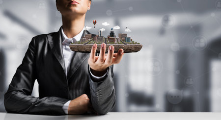 Cropped image of businessman in suit presenting in his hand modern city block with office view on background. 3D rendering.の写真素材
