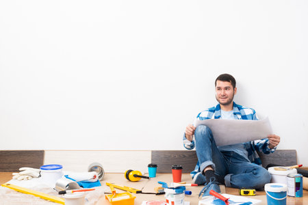 Happy guy sitting on floor with paper blueprint. Home remodeling after moving. Construction tools and materials for building. Young man wearing checkered shirt and jeans redesigning his home.の写真素材