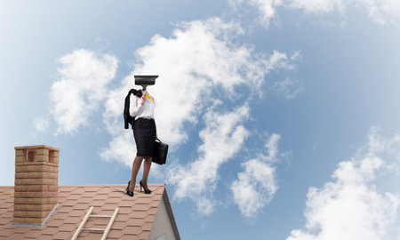 Faceless businesswoman with camera zoom instead of head standing on house roofの写真素材