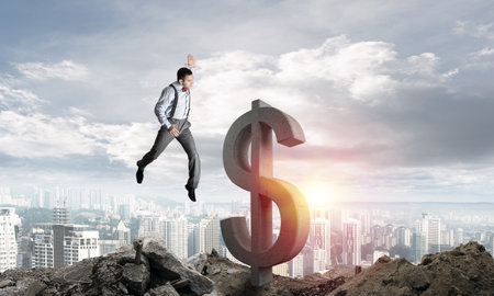 Jumping businessman in smart casual wear crashing big dollar symbol with city view on background.の写真素材