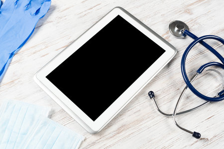 Mock up of doctors desktop with medical supplies. Tablet computer, stethoscope, disposable face mask and gloves on wooden desk. Professional healthcare technology. Medical consultation and treatment.の写真素材